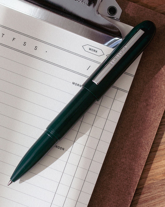 A dark green bullet pen displayed on top of a daily planning notepad and clipboard.