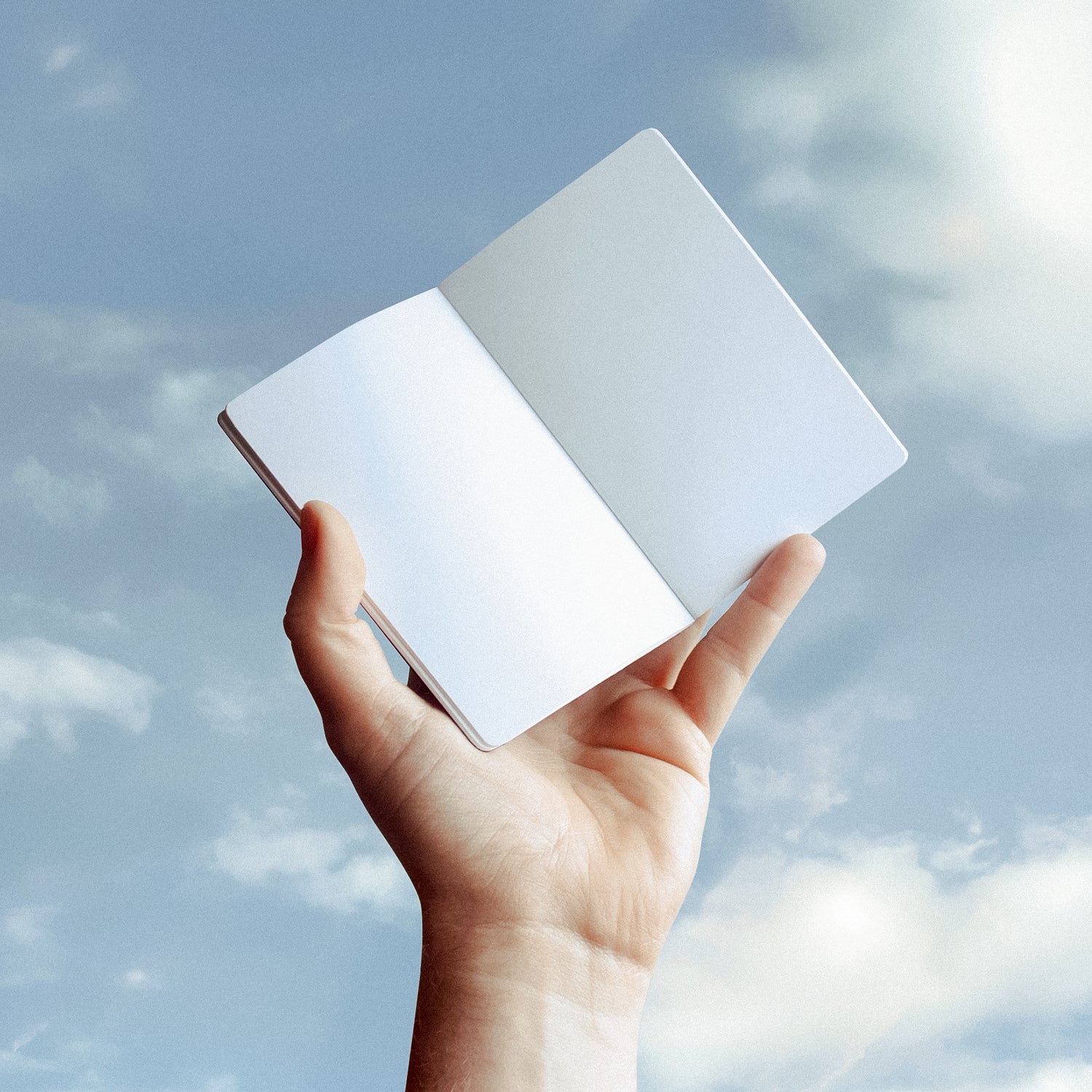 A hand holds a pocket sized notebook up against a partly cloudy sky, light blue and sunny.