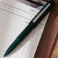 A dark green bullet pen displayed on top of a daily planning notepad and clipboard.